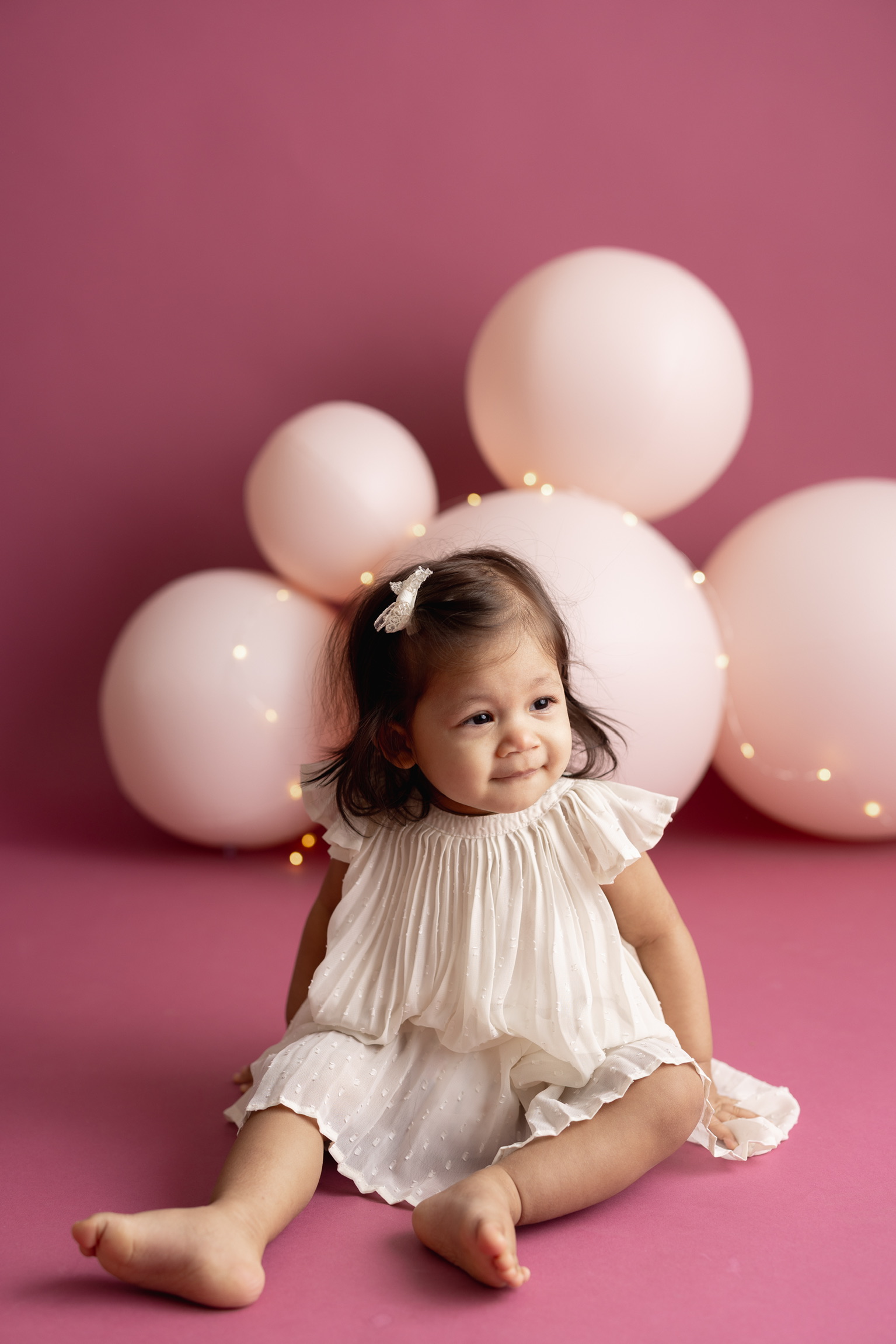 Baby girl in a cream dress and bow sitting in front of blush pink balloons on a rose backdrop