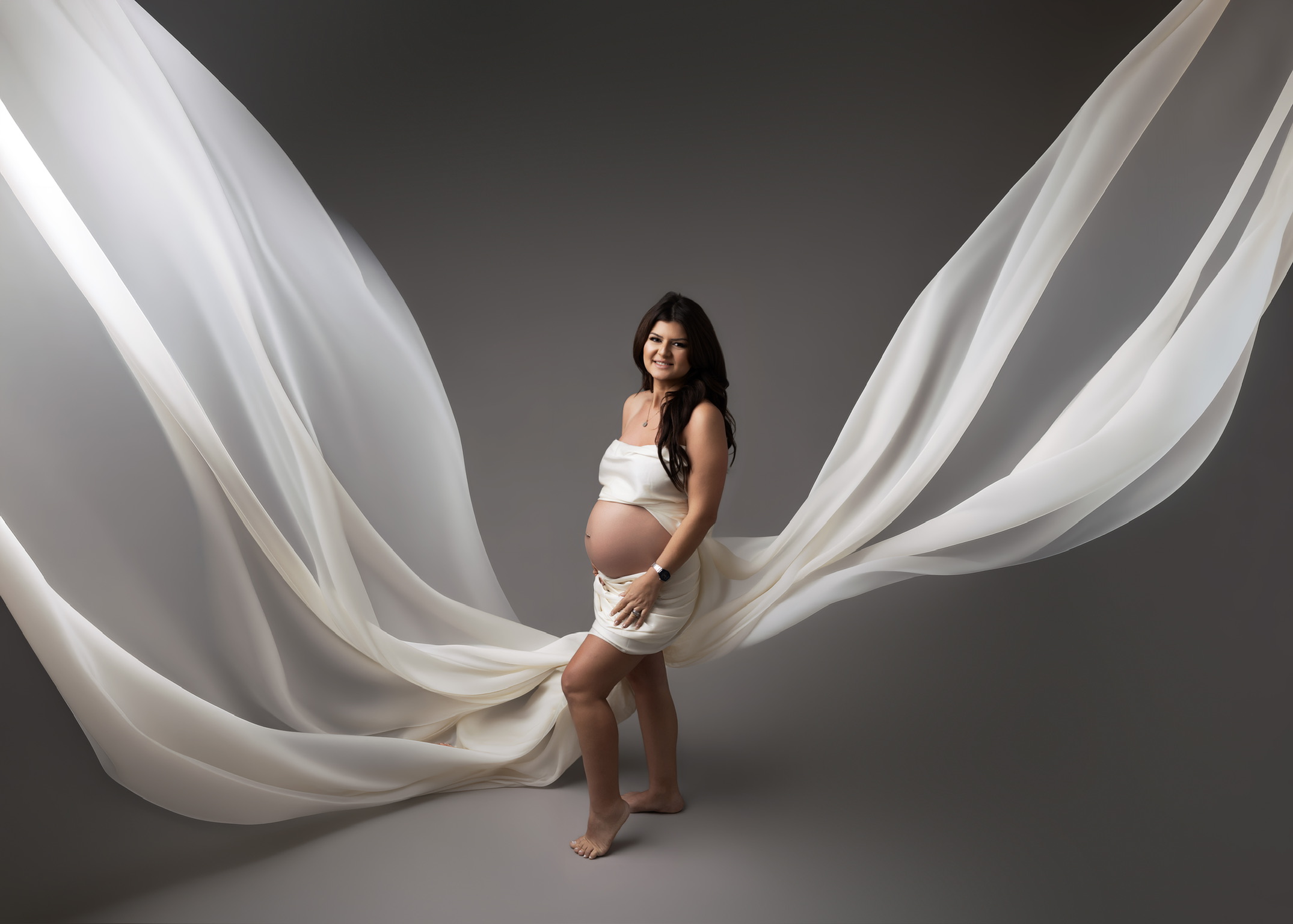 Smiling expectant mother in a white wrap as long sheer white fabric sweeps out on both sides against a gray studio backdrop
