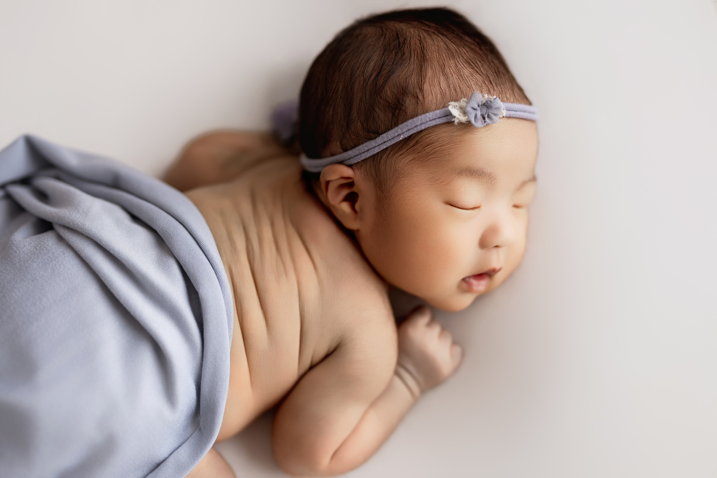 Newborn asleep on her tummy with a lilac headband and a lavender blanket