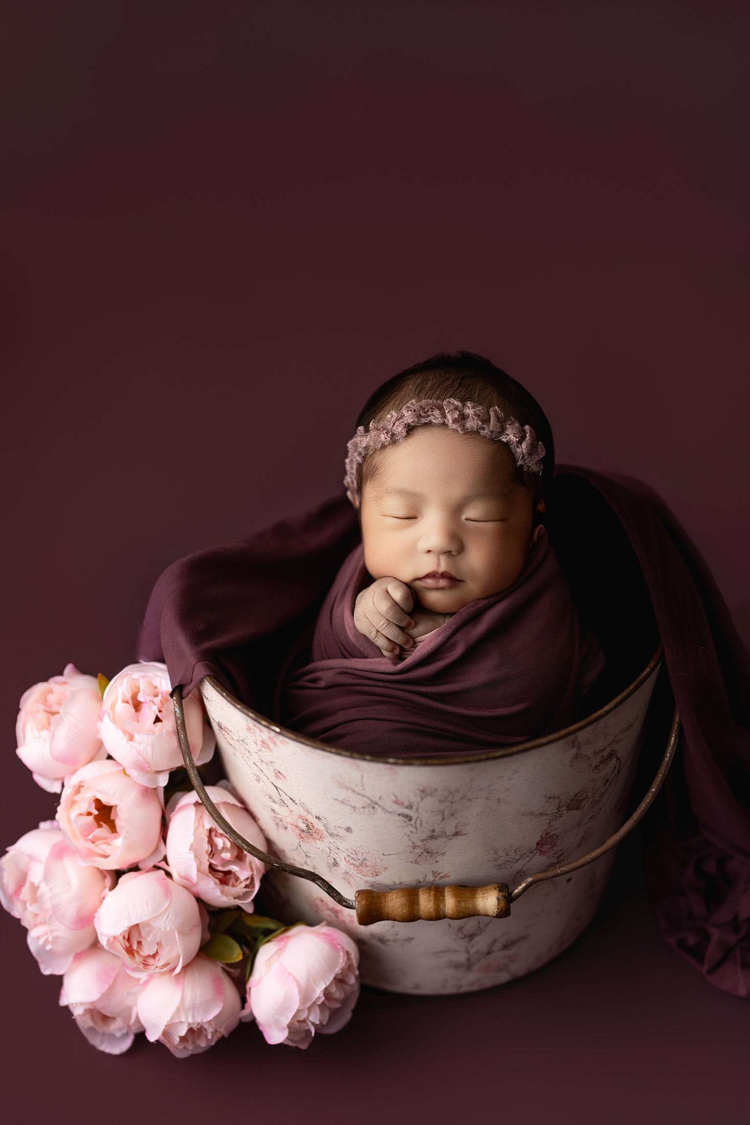 Newborn in a plum wrap and floral headband asleep in a vintage bucket beside pink peonies