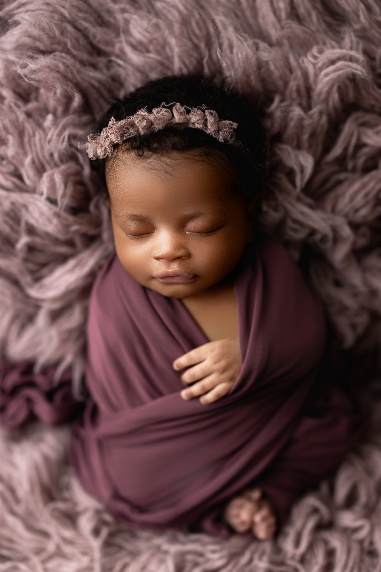 Newborn girl in a plum wrap and floral headband asleep on a soft mauve fur