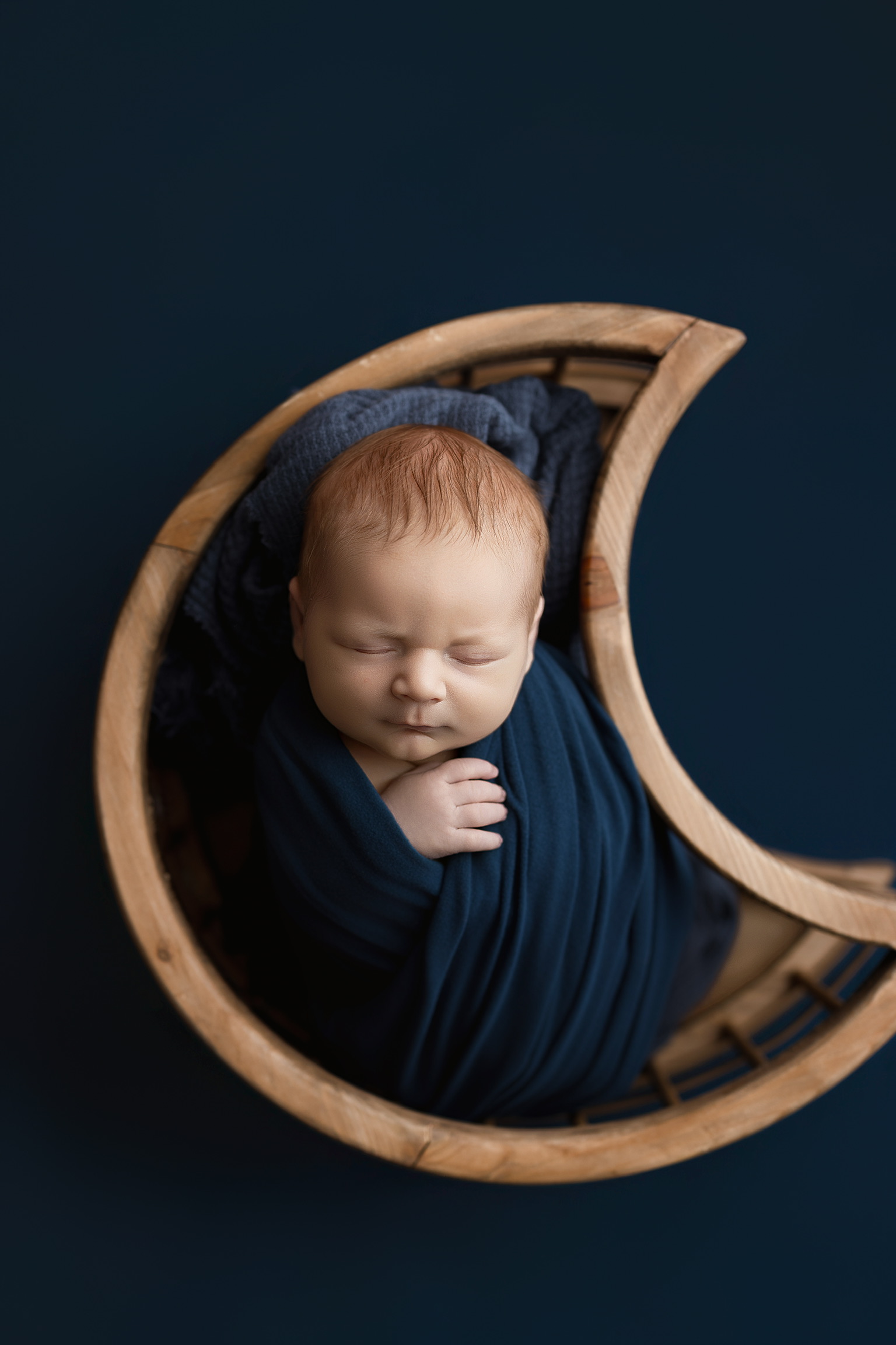 Newborn in a navy wrap sleeping inside a wooden crescent moon