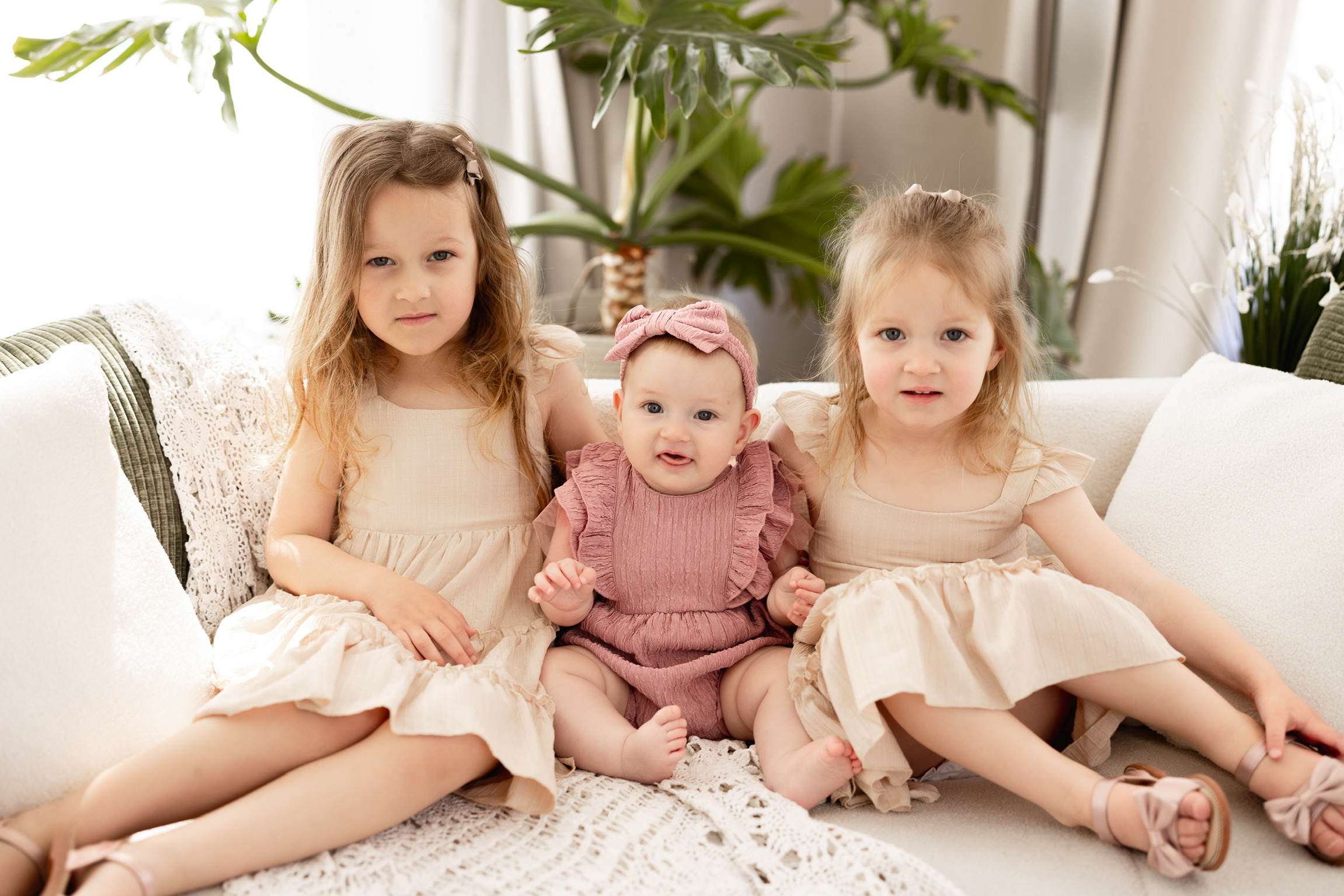 Two big sisters in cream dresses sitting on a white sofa with their baby sister between them