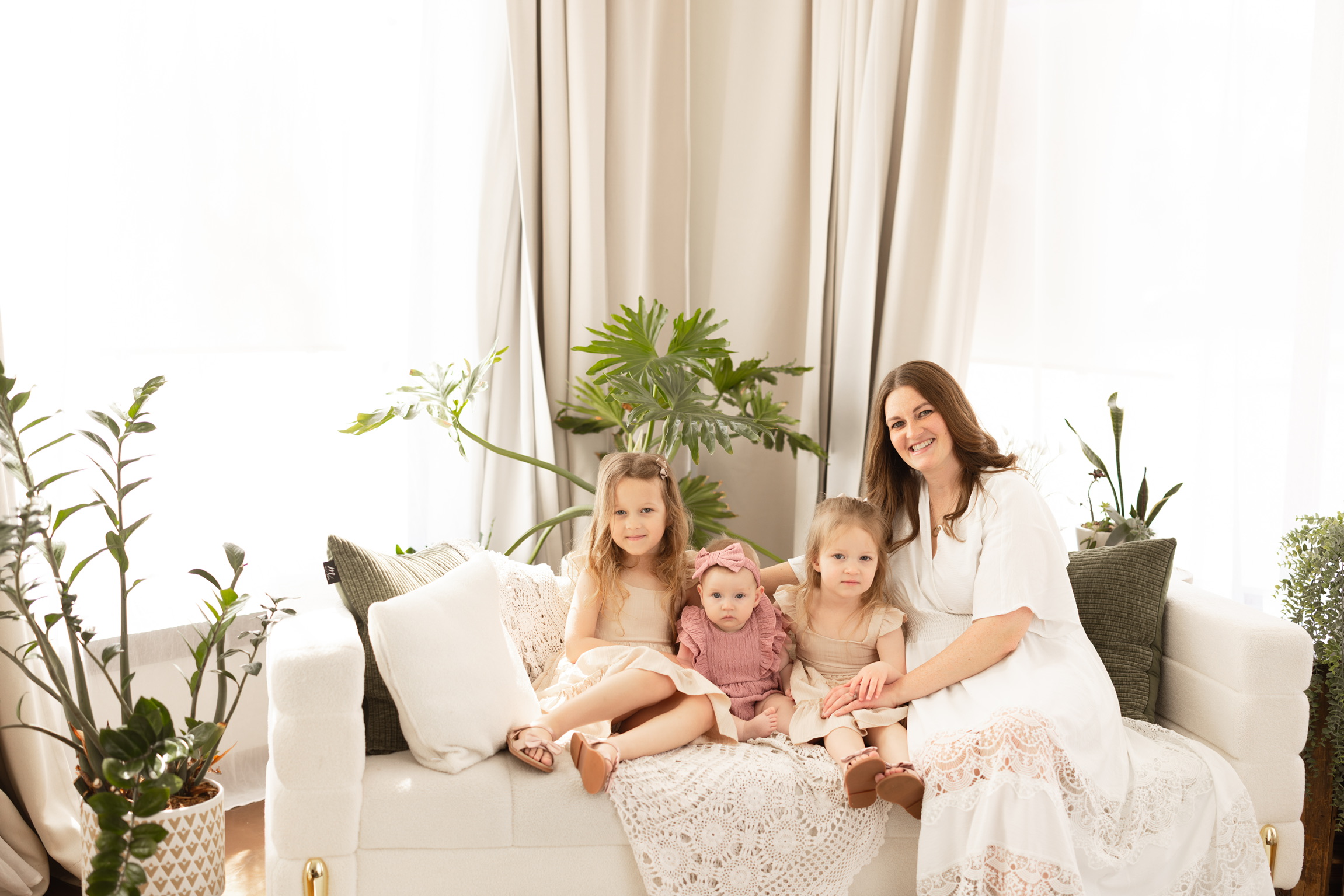 Mother relaxing with her three young daughters on a white sofa in a bright studio filled with plants