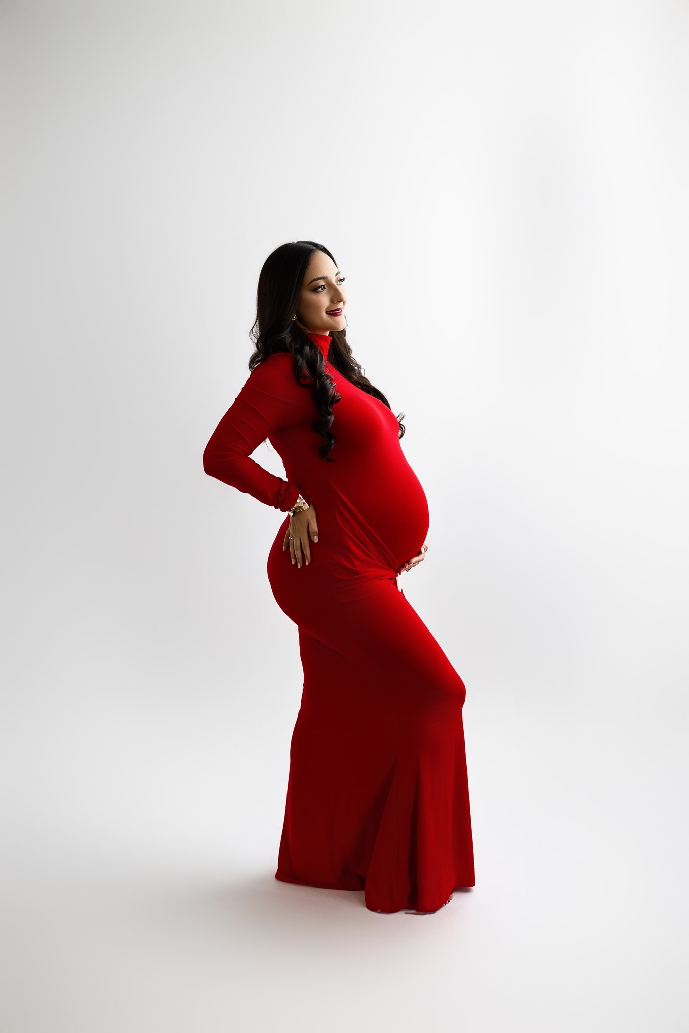 Expectant mother in a fitted red gown against a white studio backdrop