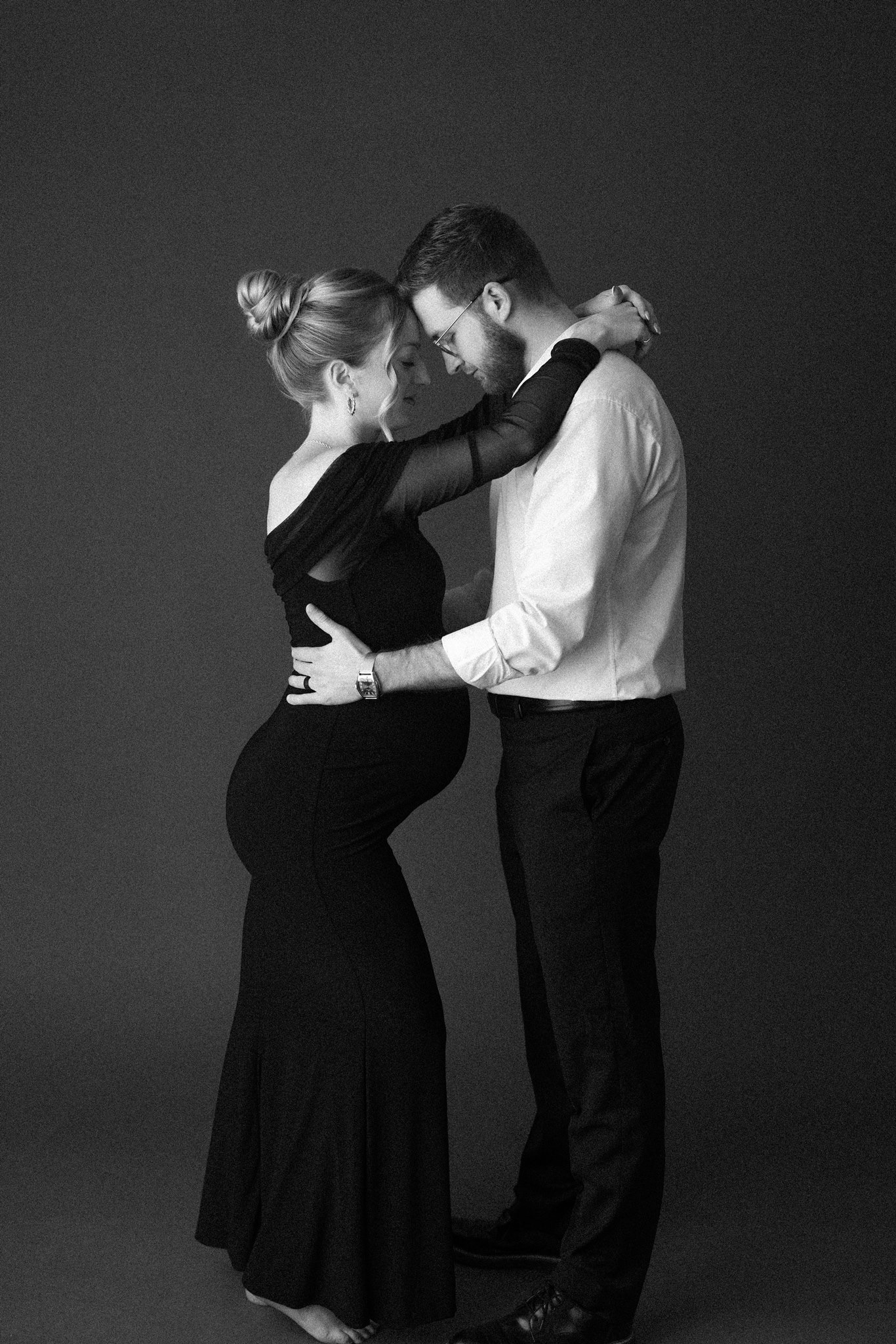 Black and white portrait of an expecting couple leaning in close, her in a fitted black gown
