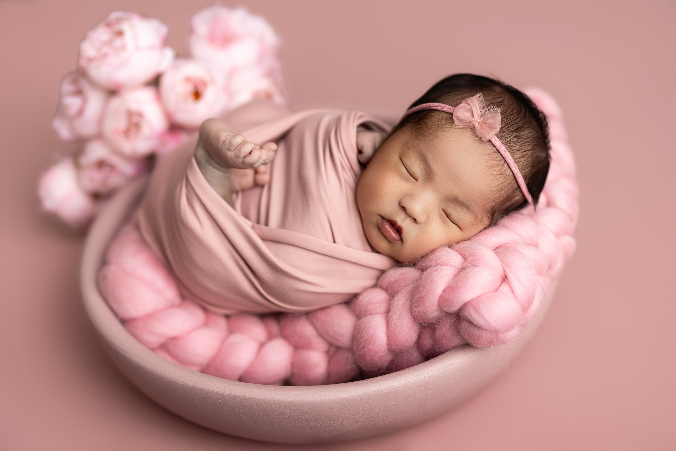 Newborn girl asleep in a blush wrap inside a pink bowl with a chunky knit blanket and roses
