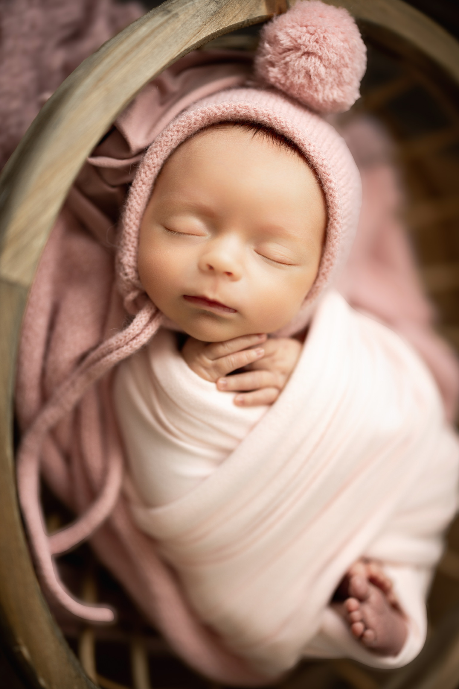 Sleeping newborn in a pink pom pom bonnet and cream wrap, tucked into a rattan basket