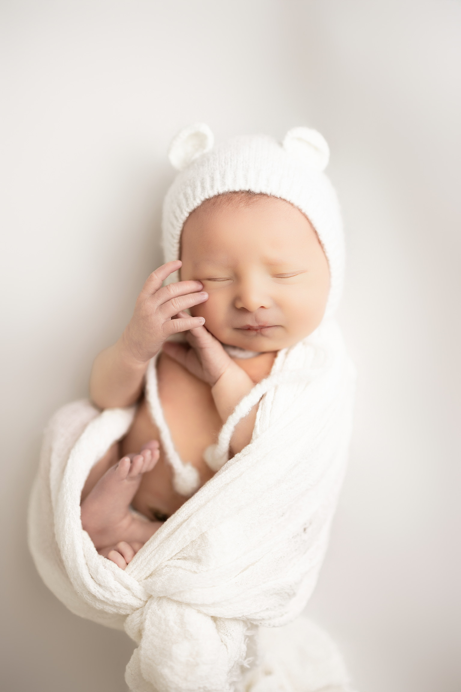 Sleeping newborn in a white knit bear bonnet with hands tucked under the chin