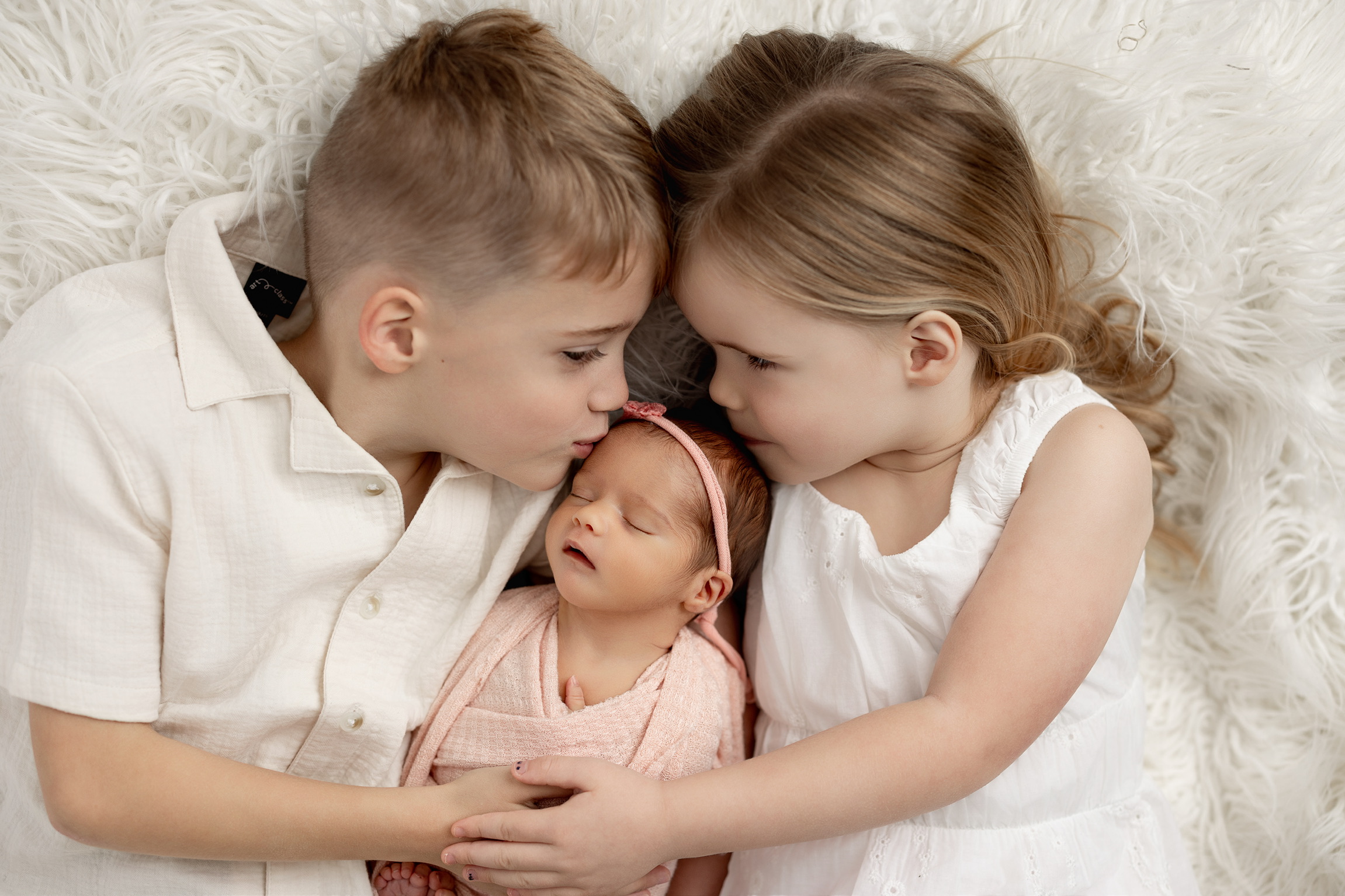 Big brother and sister leaning in to kiss their newborn sister, wrapped in blush pink on a cream rug