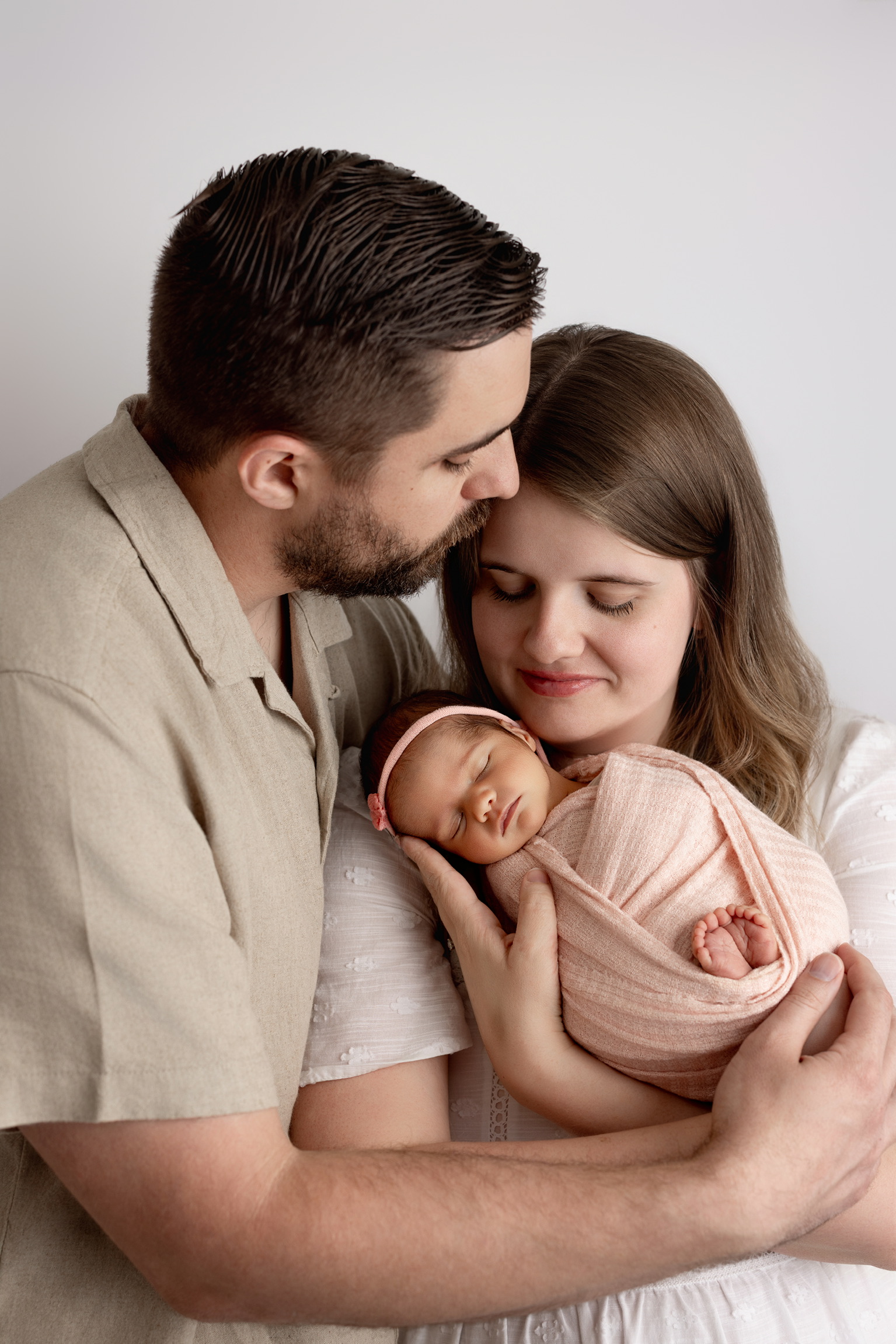 Father kissing mother on the cheek as she cradles their newborn daughter in a blush wrap