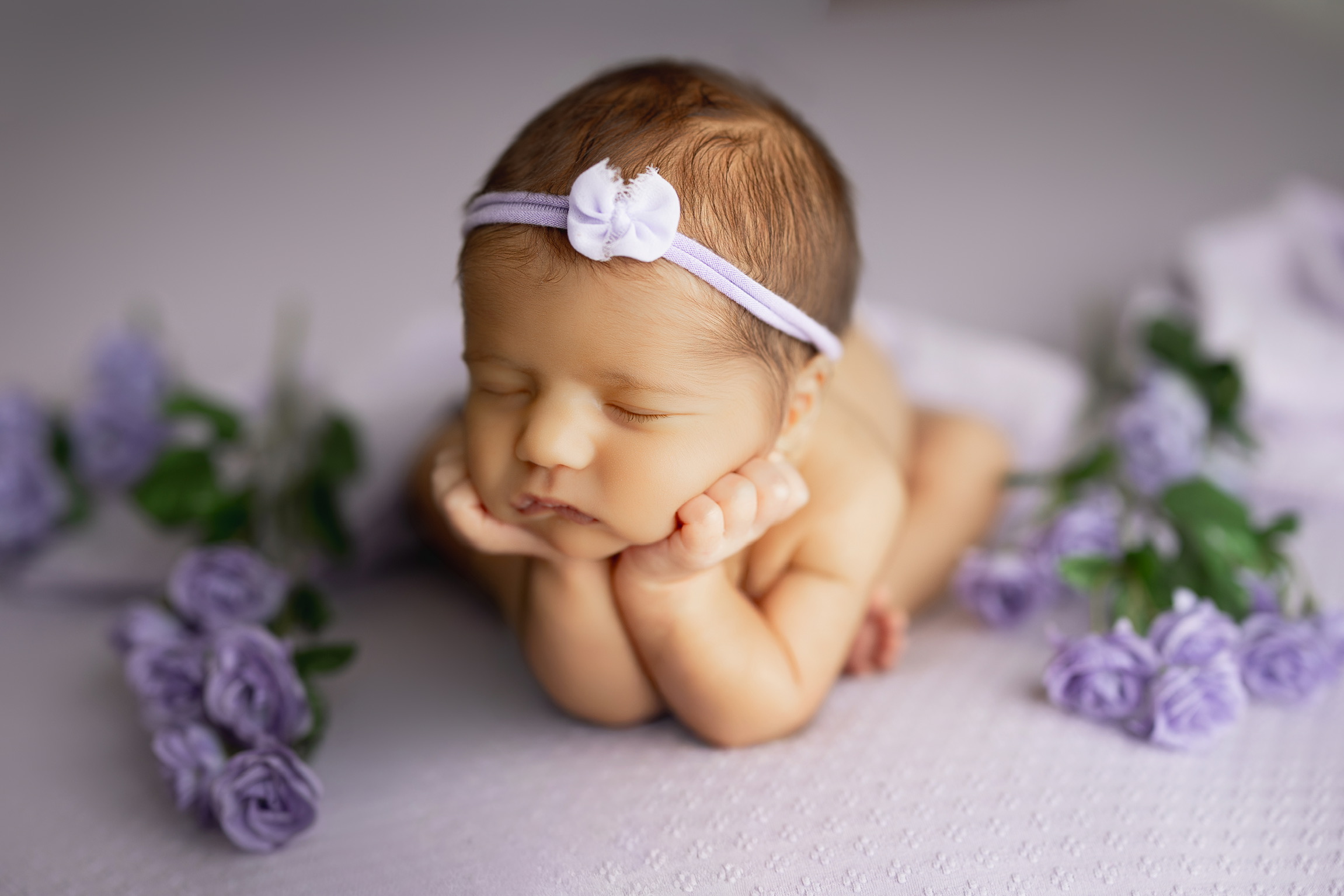 Newborn resting chin on hands on a lilac blanket, surrounded by purple flowers