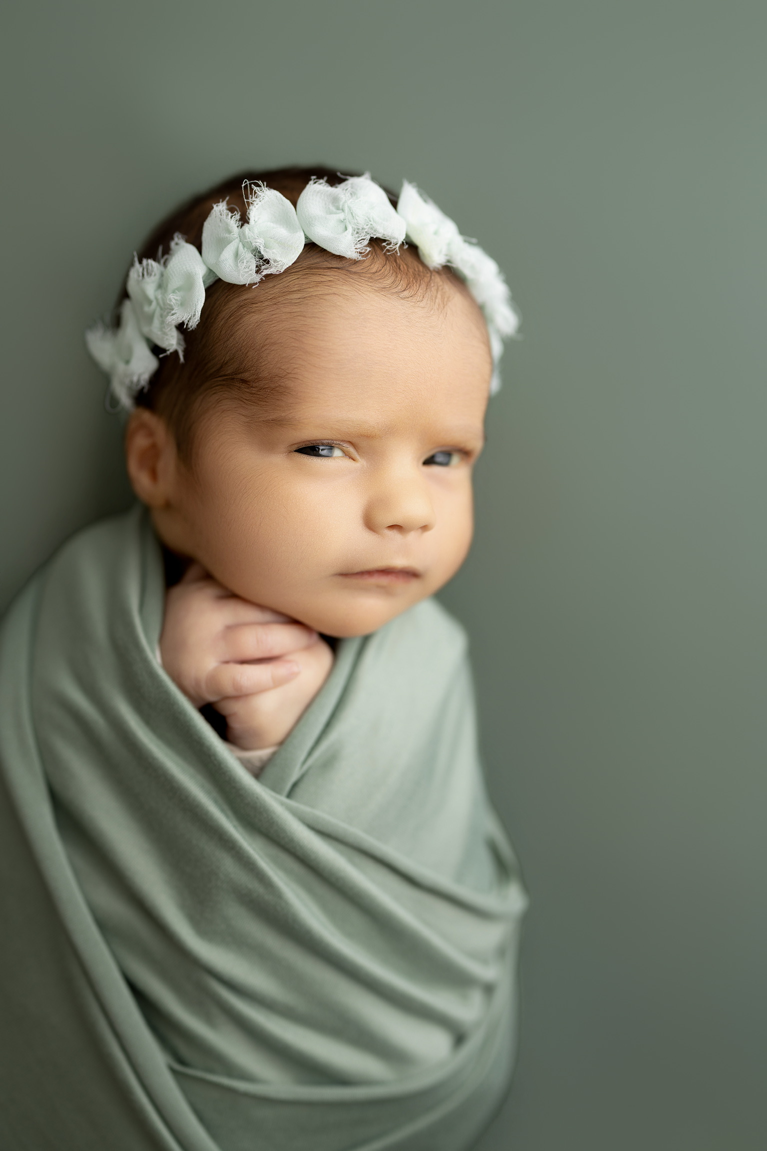 Newborn in a sage green wrap and white flower crown against a soft green backdrop