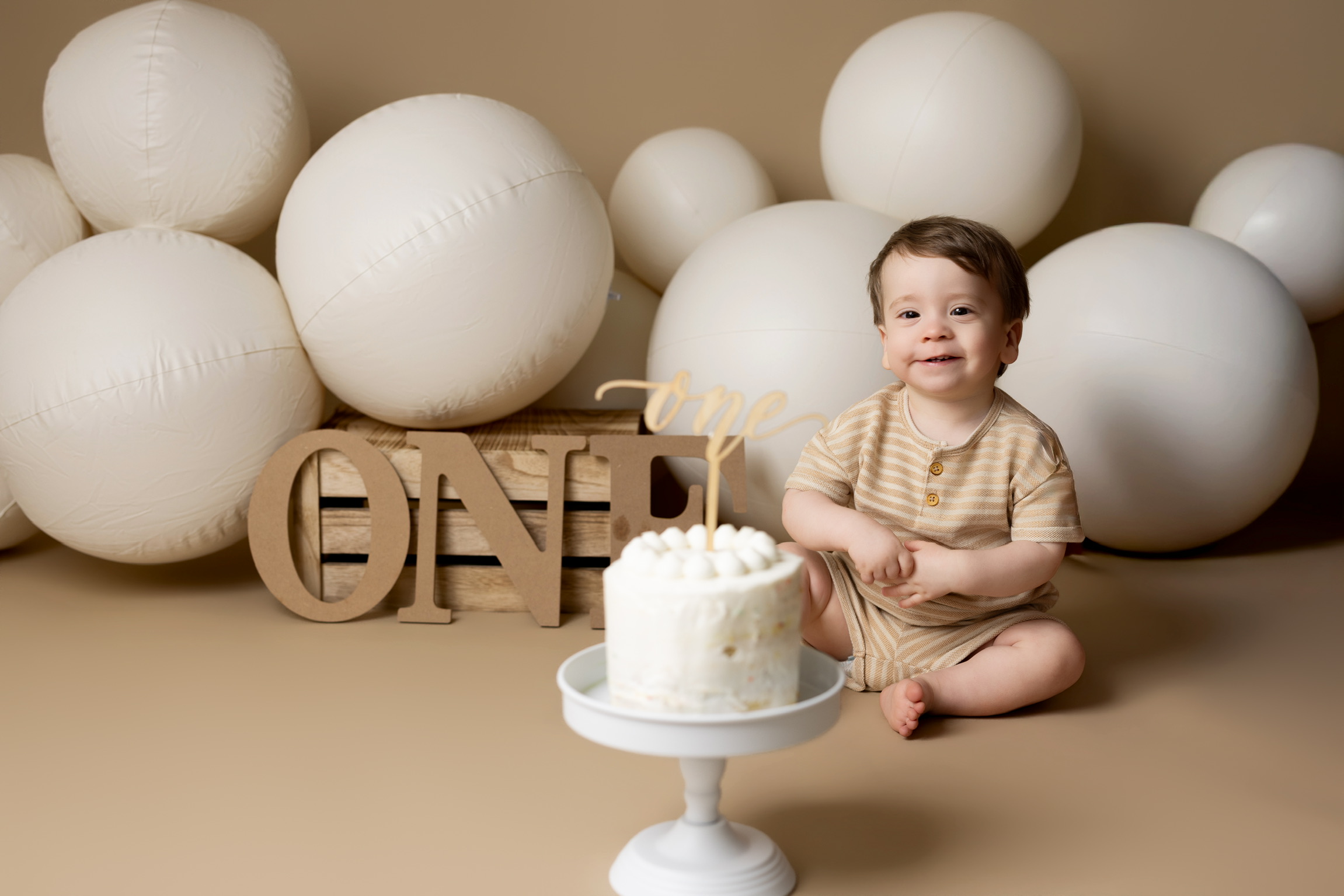 One year old boy sitting beside a small white birthday cake with wooden ONE letters and white balloons
