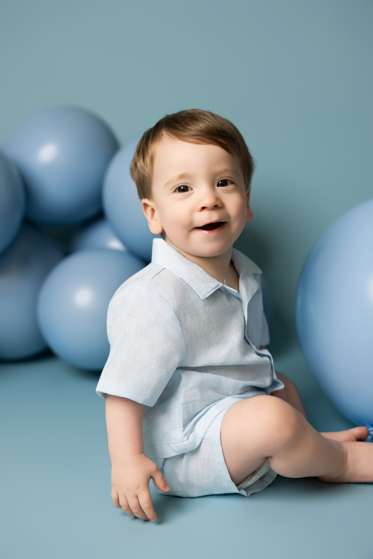 Smiling toddler boy in a light blue outfit sitting among blue balloons in the studio