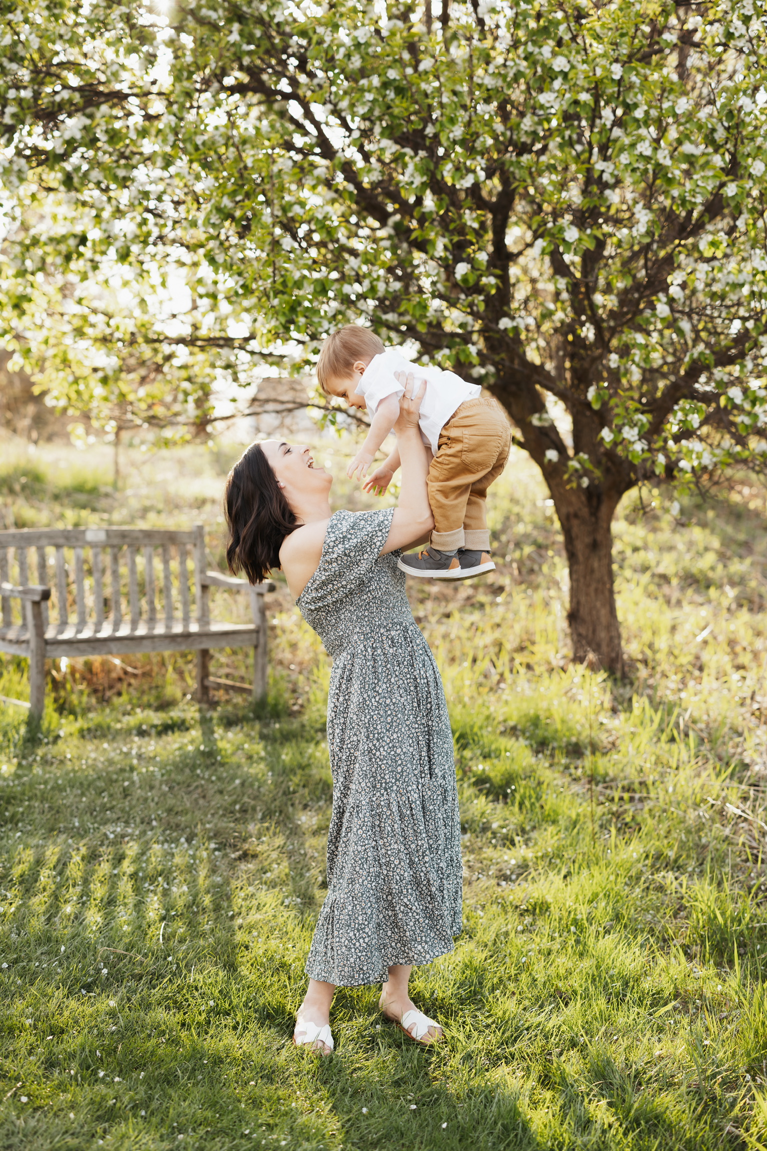 Mother in a floral dress lifting her toddler into the air under a flowering tree in a sunny field