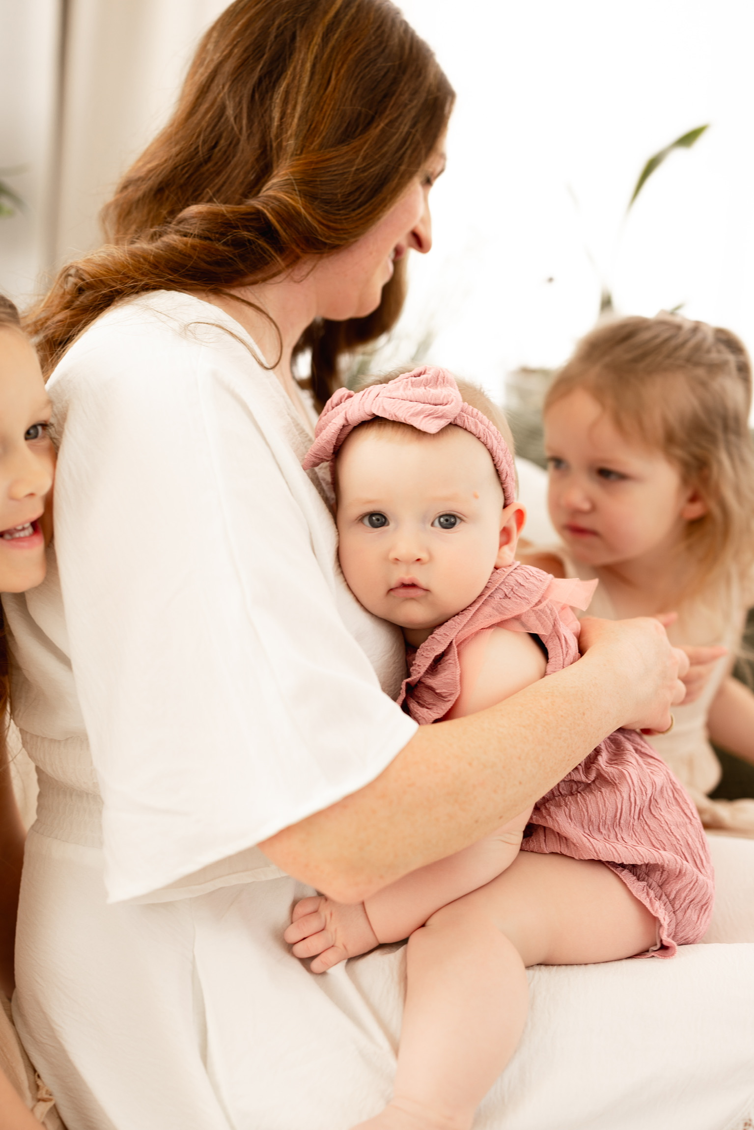Mother cuddling her baby girl in pink while big sister leans in on a white sofa