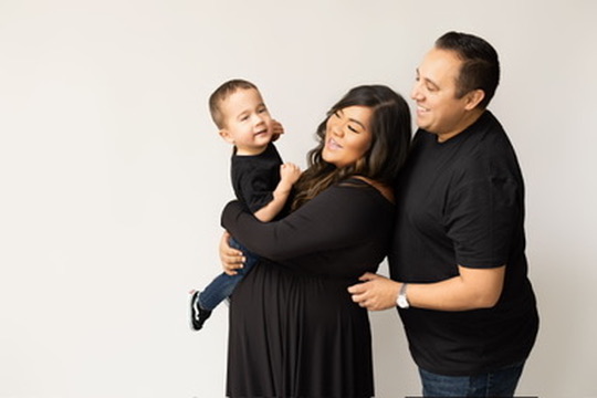 Mom and dad dressed in black laughing as they hold their toddler son against a white studio backdrop