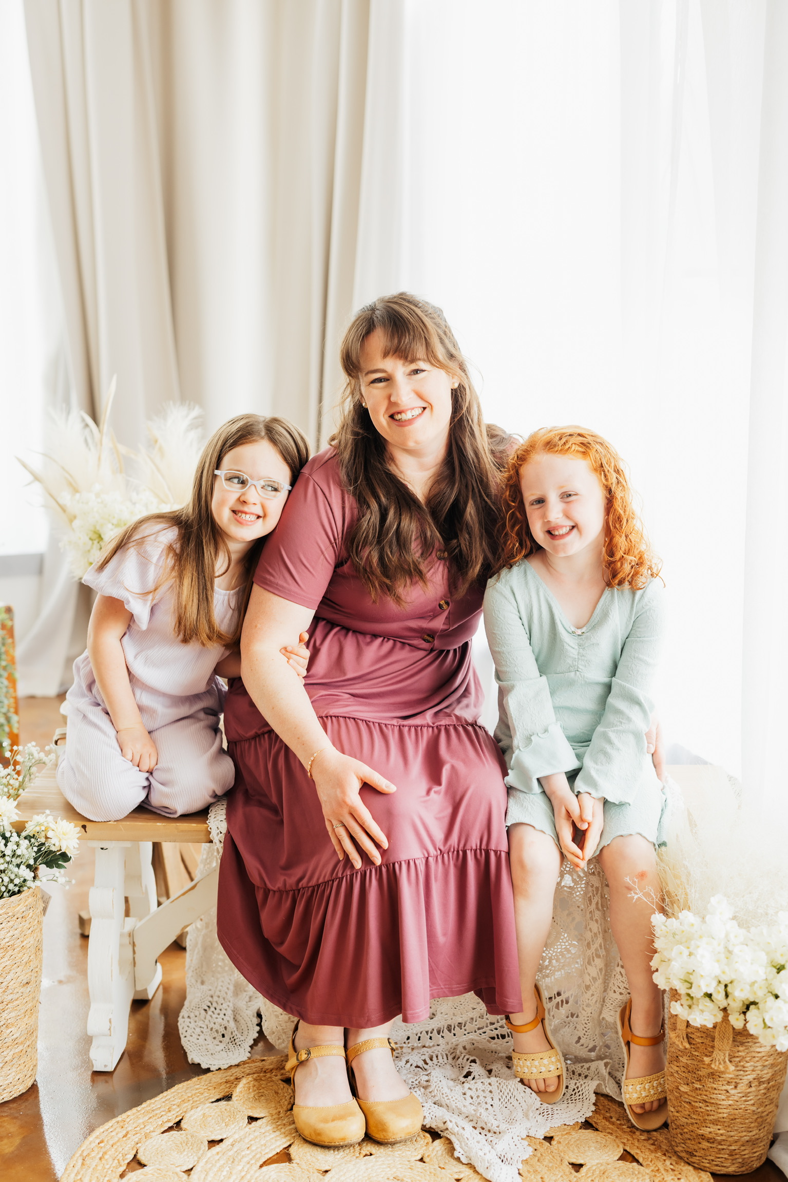 Mother seated between her two daughters in a bright studio decorated with dried flowers