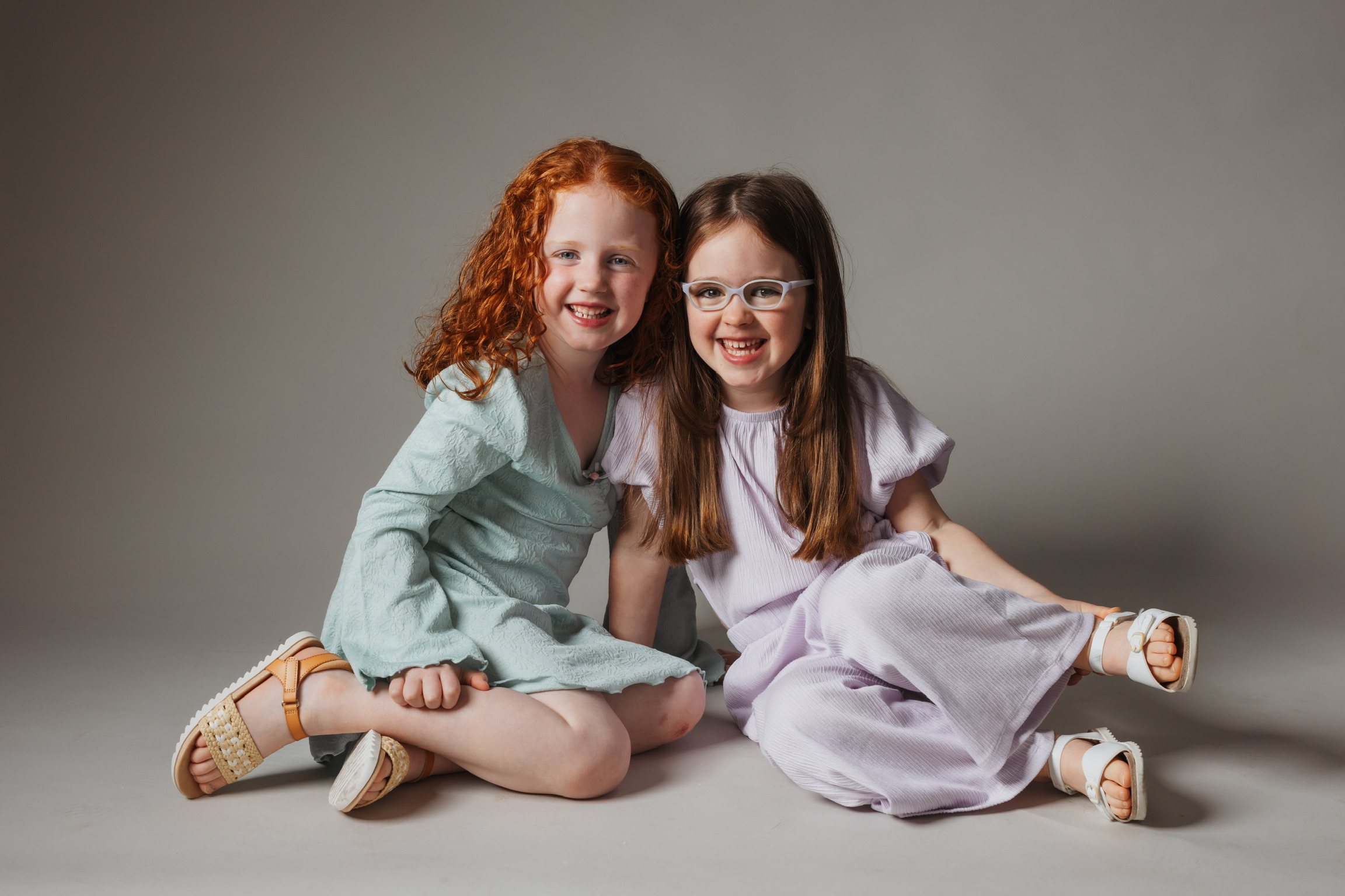 Two sisters sitting cross legged and smiling on a gray studio backdrop