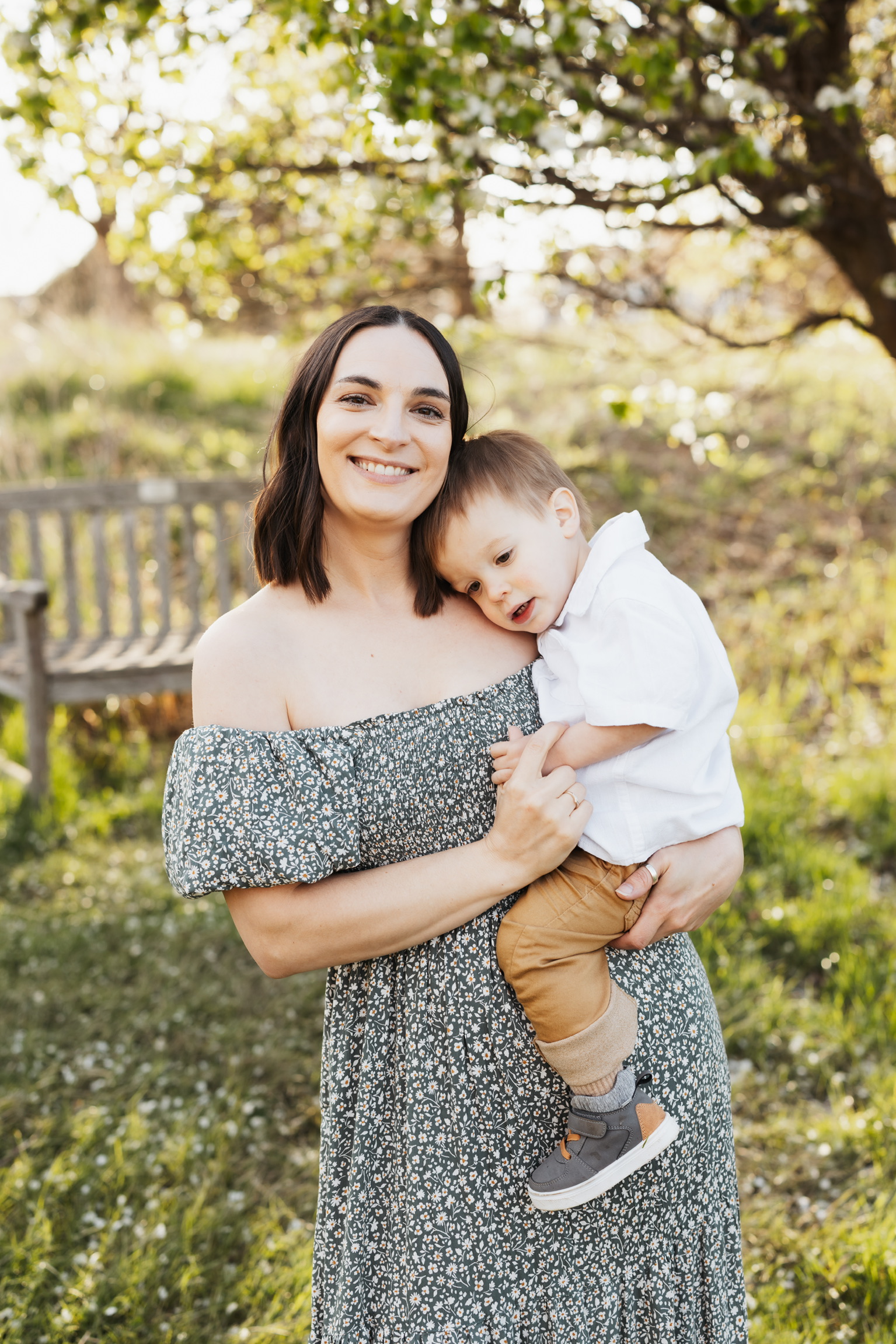 Mother in a floral dress smiling as her toddler son rests his head on her shoulder outdoors