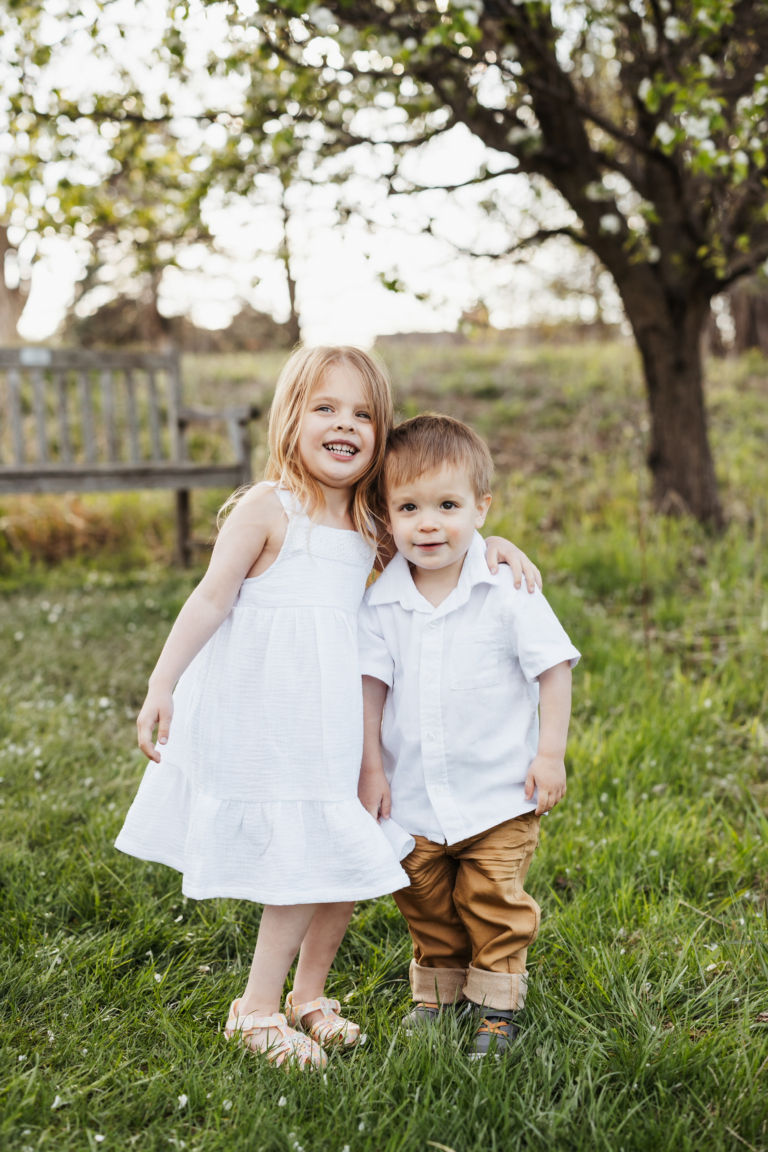 Sister in a white dress and little brother in a white shirt standing together in the grass