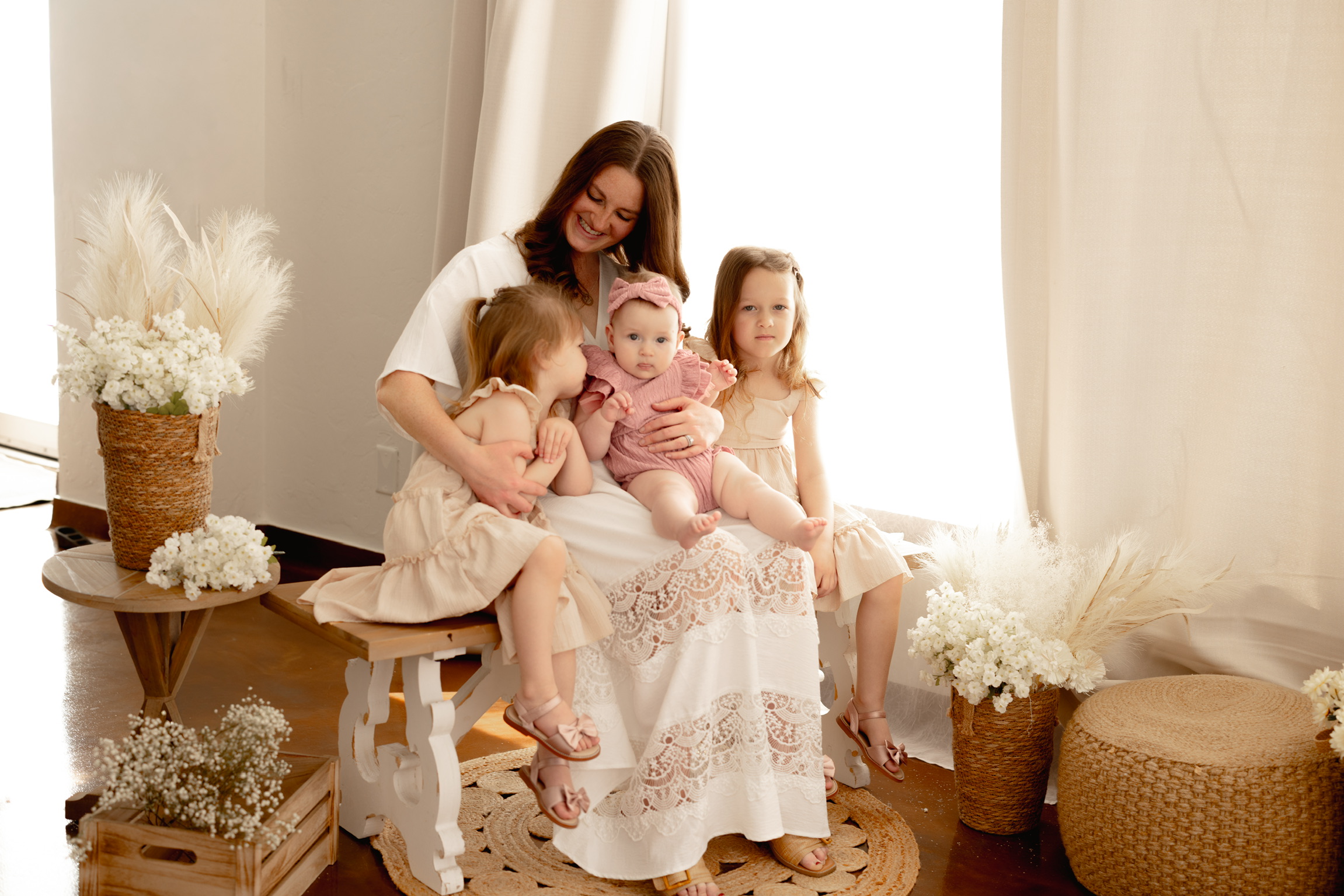 Mother holding her three daughters close in a sunlit studio styled with pampas grass